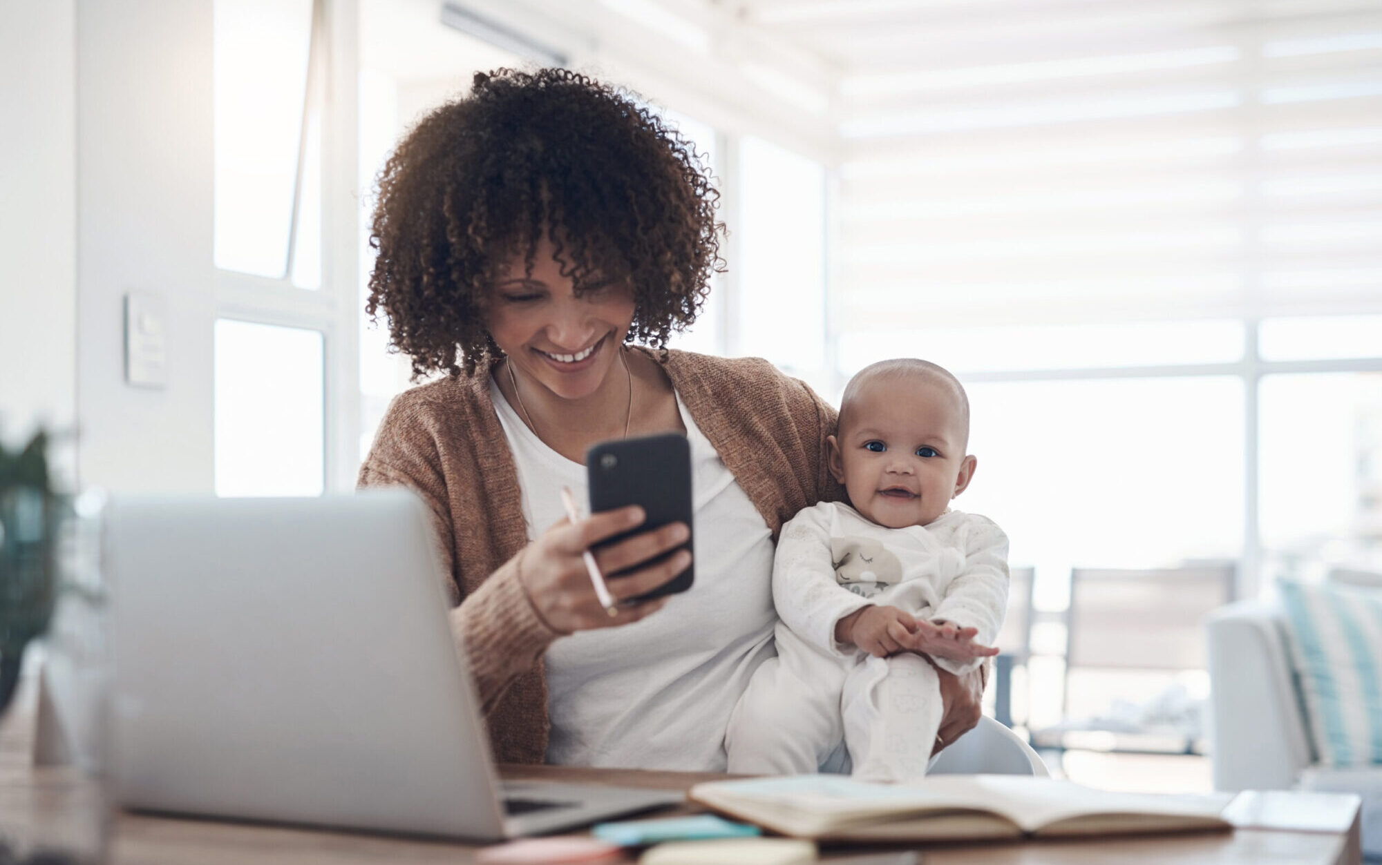 Woman with curly hair, holding baby, and booking an online appointment on her cellphone.